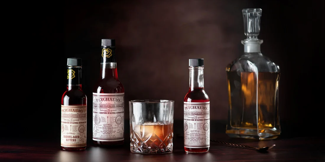 PEYCHAUD’S Aromatic and Barrel-Aged bitters beside a cocktail and decanter